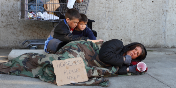 Mother laying on the ground with her children with a sign that says "Homeless, please help."